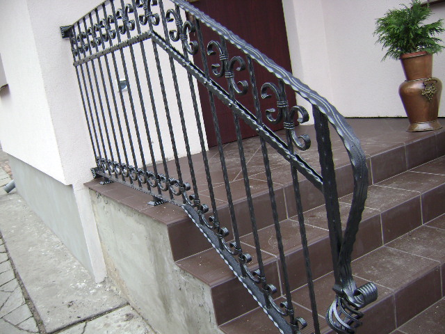 Kuta, metalowa balustrada schodowa z ozdobnymi elementami, prowadząca do drzwi wejściowych. Widoczny fragment elewacji budynku i donica z rośliną na schodach.